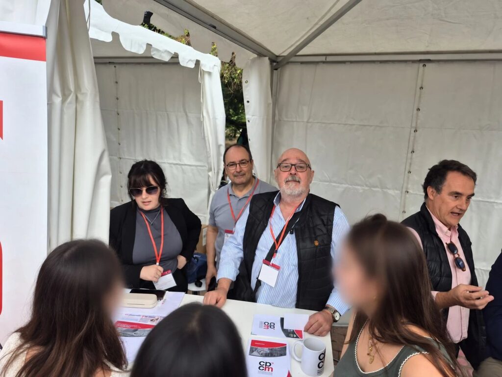 Representantes del CPCM atienden desde la mesa de la carpa a un grupo de estudiantes, con folletos del Colegio sobre la mesa.